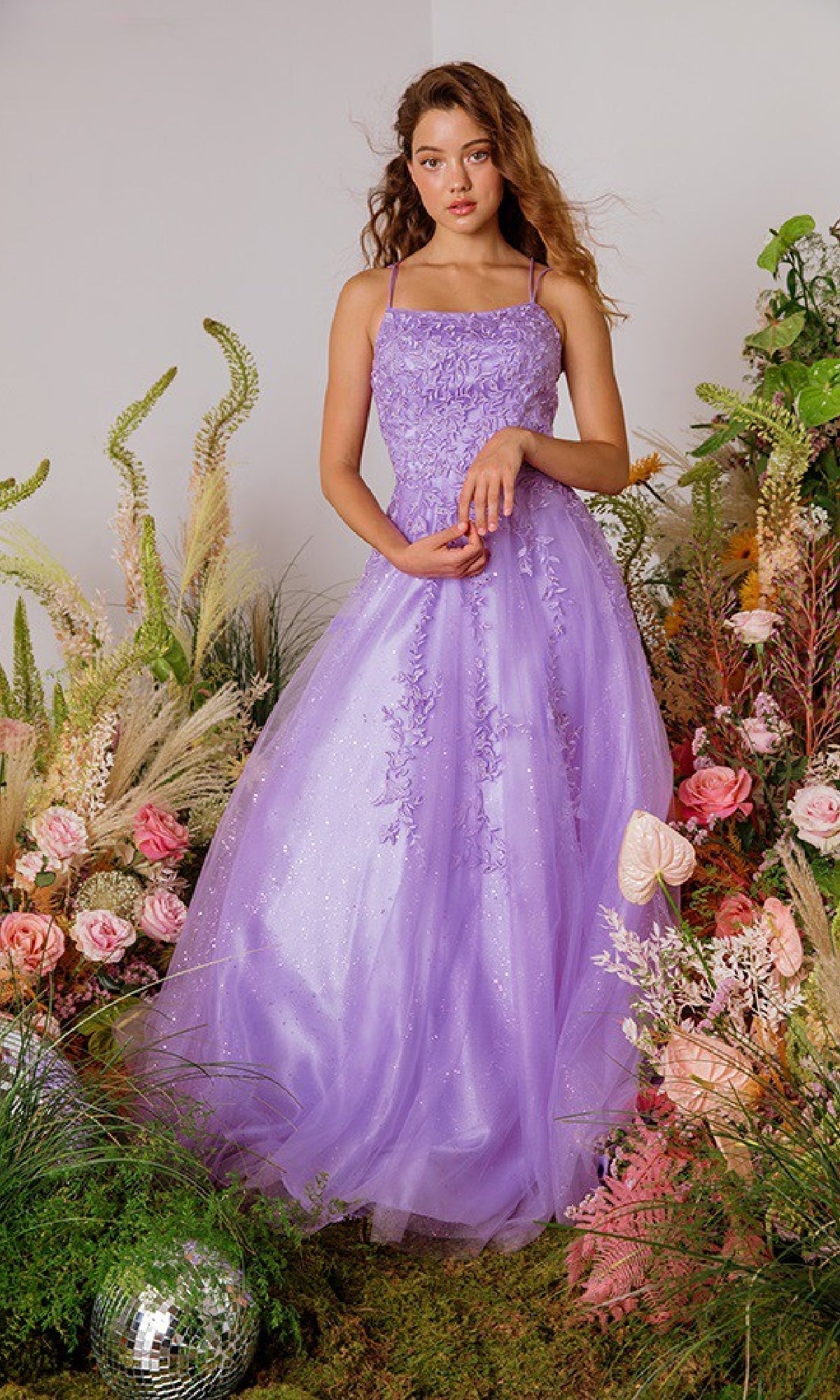 A young woman in the Lace-Up Long Lace Prom Ball Gown: Eureka CC0240 stands among pink flowers, grasses, and foliage against a neutral backdrop.
